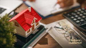 Casa em miniatura sobre documentos, com calculadora e itens de análise financeira ao fundo.