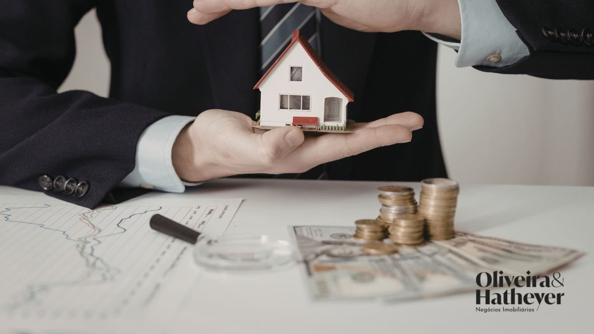 Profissional protegendo uma casa em miniatura sobre moedas e documentos financeiros.
